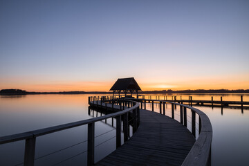 Obraz premium Swinged Pier construction and shelter with thatched roof at beautiful colorful sunrise under clear sky at Lake Hemmelsdorf, Schleswig-Holstein, Northern Germany