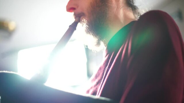 A bearded man plays the old pipe. He blows air into the tube, makes a sound. Hands pinches holes, makes a melody.