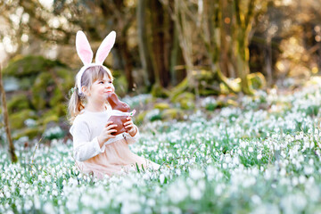 Adorable little girl with Easter bunny ears eating chocholate figure in spring forest on sunny day,...