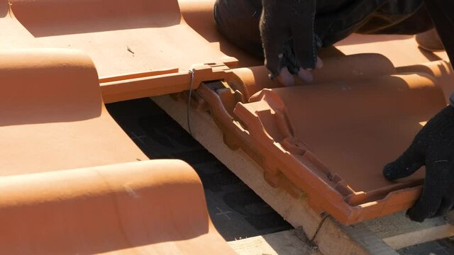 Closeup of worker hands installing yellow ceramic roofing tiles mounted on wooden boards covering residential building roof under construction.