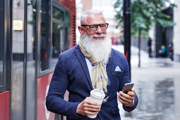 Business man using cellphone outdoors while waiting for the bus. Portrait of funny attractive cheerful pensioner hold hand hot takeout coffee beverage and smartphone. Transport and job concept.