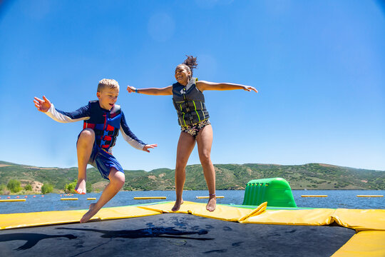 Two happy diverse kids jumping and playing on a inflatable water bounce house on a lake. Jumping on a inflatable trampoline on a summer day is super fun!
