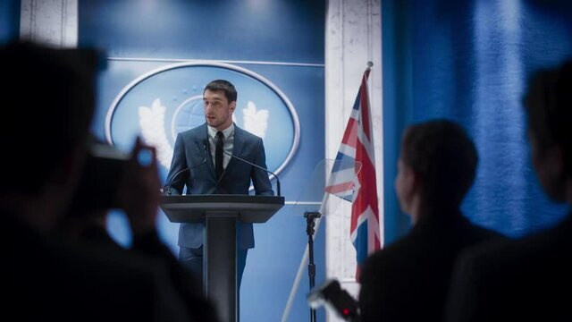 Young Organization Representative Speaking at Press Conference in Government Building. Press Officer Delivering a Speech at Summit. Minister Speaking to Congress. Backdrop with Great Britain Flags.