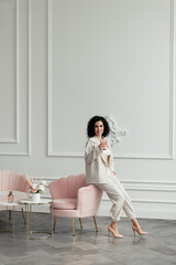 Woman with dark curly hair sits on an armchair holding a glass and looks at the camera