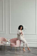 Woman with dark curly hair sits on an armchair holding a glass against a wall background