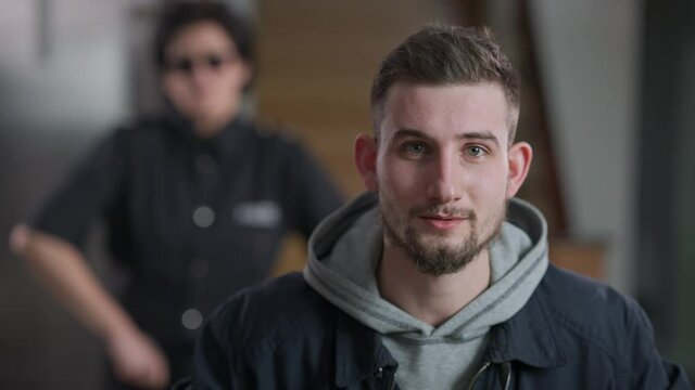 Young brunette Caucasian man with green eyes looking at camera talking as blurred police officer shaking head at background. Portrait of unfortunate burglar persuading innocence.