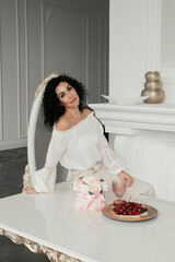 A woman in a white blouse sits on a table next to a plate of cherries and a bouquet of flowers