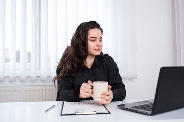 Smiling girl looking at the laptop while making a coffee break at her office.