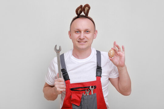 Easter holiday, celebration concept. funny smiling engineer worker or mechanic is wearing rabbit ears on head, holding wrenches and painted egg white background. celebrating orthodox day - Powered by Adobe