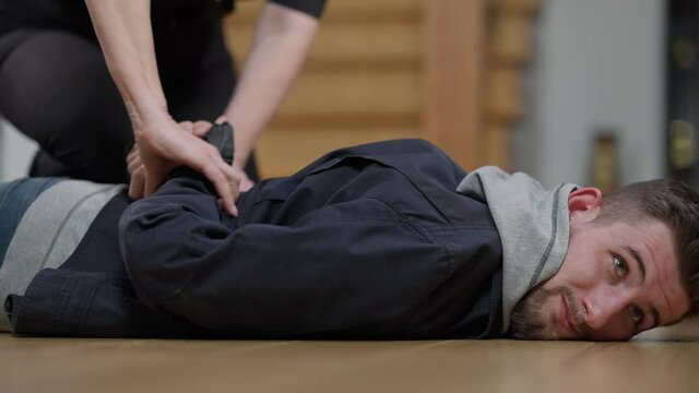 Caught young criminal lying on floor in police lights as unrecognizable policewoman putting his hands behind back. Unsuccessful thief or murderer arrested by police officer indoors at crime scene.