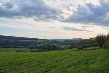 Landscape. Green field. Grass. Tractor. Bushes. Trees. Hills. There's a village in the distance. Blue sky with white and blue-gray clouds.