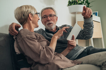 Embracing senior couple using digital tablet together at home. Happy senior couple sitting at couch at living room, talking, smiling, showing and looking in white portable computer