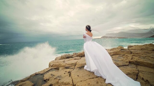 Amazing view a young bride in a white wedding dress and veil standing on a rock against the backdrop of sea or ocean and crashing waves Attractive young woman in nature on a background of landscape