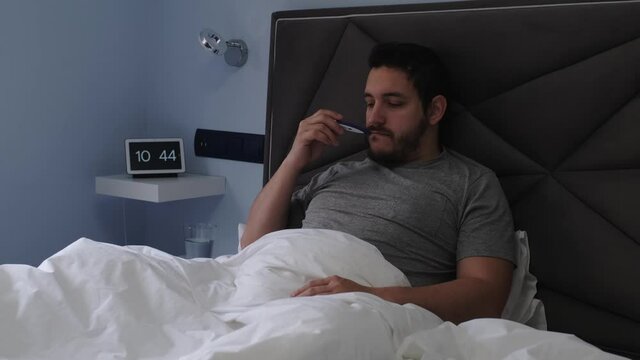 Young latin man ill in bed measuring temperature with a thermometer and taking a pill and a glass of water. Feeling sick concept. 4k video.