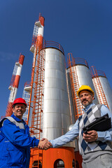 Businessperson and Worker in Uniform Shaking Hands Against Power Plant Chimneys and Storage Tanks