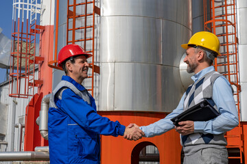 Worker in Uniform and Businessperson Shaking Hands Against Power Plant or Oil Refinery Storage Tanks
