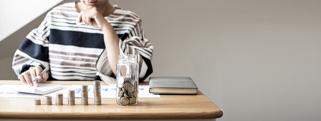 Piles of coins and a glass jar are placed on the desk of a woman sitting in keeping a savings...