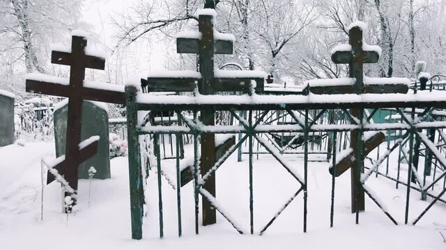 View of the cemetery with the wooden crosses and fences among falling snowflakes in a slow motion. There are crucifixes and tombs at a frosty day on the trees background. Concept of death and religion