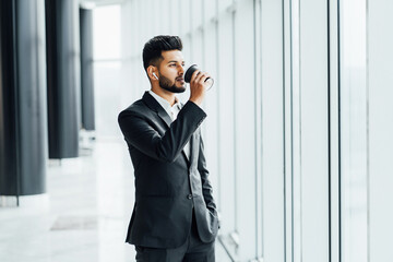 Modern Indian man in a black suit drinks his coffee in the middle of a modern building, office worker, boss, businessman