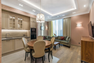 Spacious kitchen with the large lounge area with a soft velvet olive sofa. Neoclassic beige kitchen with built-in appliances. The round  table with soft chairs standing in the center of the kitchen