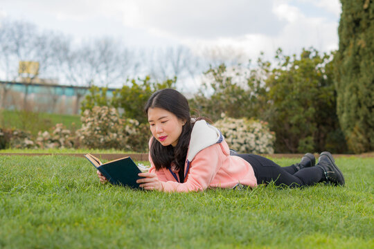 young Asian woman enjoying novel on grass - lifestyle portrait of young happy and pretty Japanese girl reading a book at beautiful city park in reading and studying concept