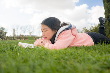 young Asian woman enjoying novel on grass - lifestyle portrait of young happy and pretty Korean girl reading a book at beautiful city park in reading and studying concept