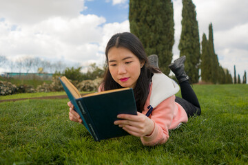 Naklejka na ściany i meble young Asian woman enjoying novel on grass - lifestyle portrait of young happy and pretty Korean girl reading a book at beautiful city park in reading and studying concept