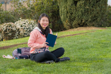 Naklejka na ściany i meble young Asian woman enjoying novel on grass - lifestyle portrait of young happy and pretty Korean girl reading a book at beautiful city park in reading and studying concept