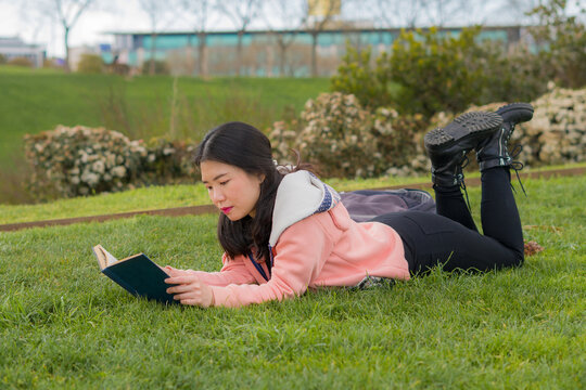 young Asian woman enjoying novel on grass - lifestyle portrait of young happy and pretty Korean girl reading a book at beautiful city park in reading and studying concept