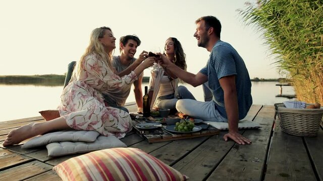 Group of friends having fun on picnic near a lake, sitting on pier eating and drinking wine.
