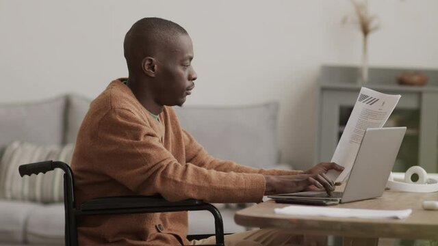 Medium shot of young handsome African disabled businessman sitting on wheelchair at desk in living room with document in one of his hands and typing on laptop keyboard with the other one