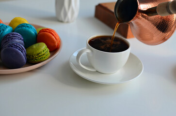Pouring coffee into a cup from a turkish coffee pot, multicolored macaroons on white background