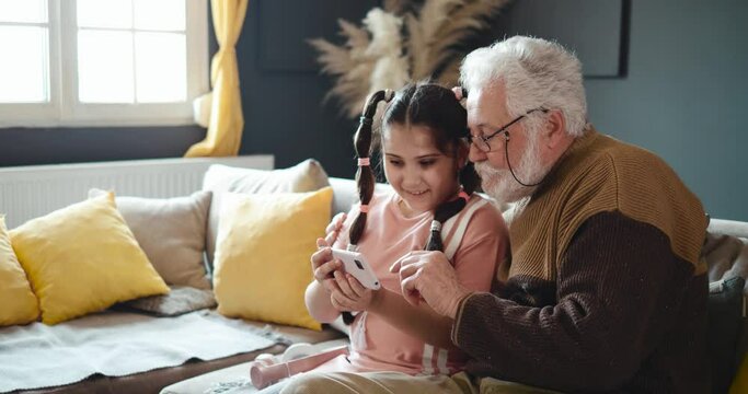 Little girl shows her grandfather how to use a smartphone. Senior man with a grandson at home uses the phone