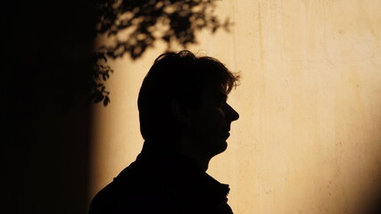 silhouette of a man in profile, who follows someone along the road, against the background of a beige wall at sunset 