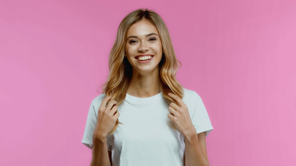 happy young woman touching hair and smiling isolated on pink