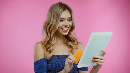 cheerful woman holding credit card and digital tablet isolated on pink