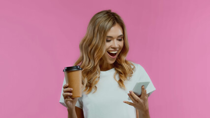 amazed woman holding paper cup and using smartphone isolated on pink