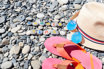 Summer travel accessories as sun hat, round mirror sun glasses, pink flip flops lie on grey round sea stones with inscription summertime. Preparations for spending vacations on sea.