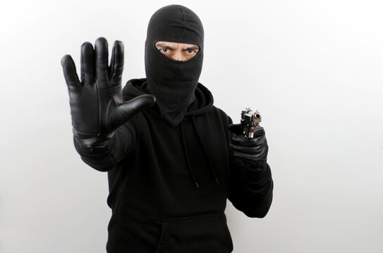 Man in black mask holding pistol and with hand doing negative stop sign, over white background. Crime concept