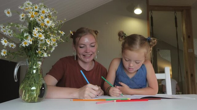 Smiling mother and her daughter are drawing with colored pencils in an album. Family cute blondes are having a good time together. Concept: motherhood and mother's day