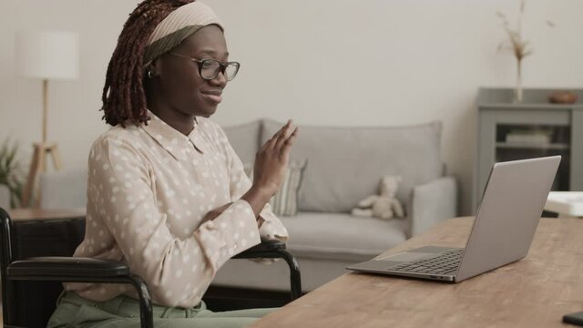 Medium shot of young attractive African disabled woman sitting on wheelchair at desk in living room and typing on laptop keyboard