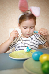 little cute blue-eyed girl in bunny ears paints Easter eggs