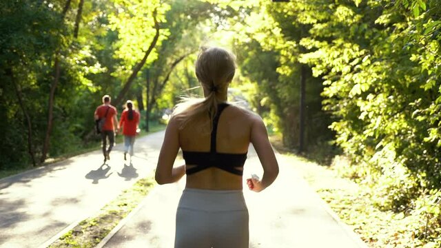 Blonde woman with ponytail wearing sexy sportswear jogging in sunny park, unrecognizable people walking beside. Following shot of fit female exercising outside in summer. Concept of sport