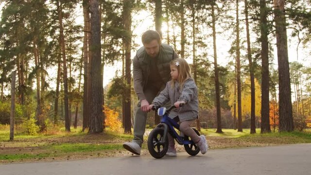 Tracking shot of father helping little 5-year-old daughter riding balance bicycle along asphalt path in park on sunny autumn day