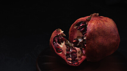 ripe red shrunken notched pomegranate with slice on a clay plate on a black background. Moody still life with copy space