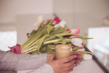 Cappuccino in a glass glass. The girl holds in her hands a cup of coffee with a pattern with a bouquet of tulips on the table.
