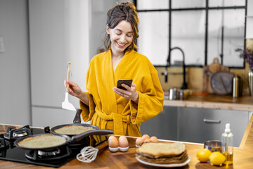 Happy woman dressed in bathrobe cooks pancakes for breakfast, looks on phone while standing near the hob in the modern kitchen at home