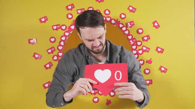 man on an isolated yellow background holds a sign in the form of a heart, he is upset that he has zero likes on social networks.