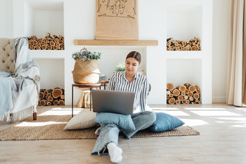 Young caucasian woman working on laptop in cozy living room, sitting on the floor with pillows.