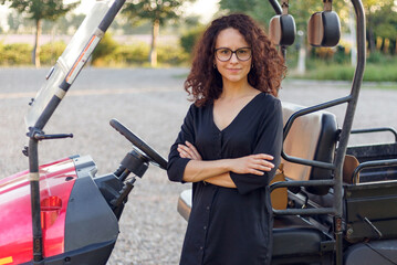 Frontal portrait of a cheerful woman with crossed hands poses near vehicle outside.